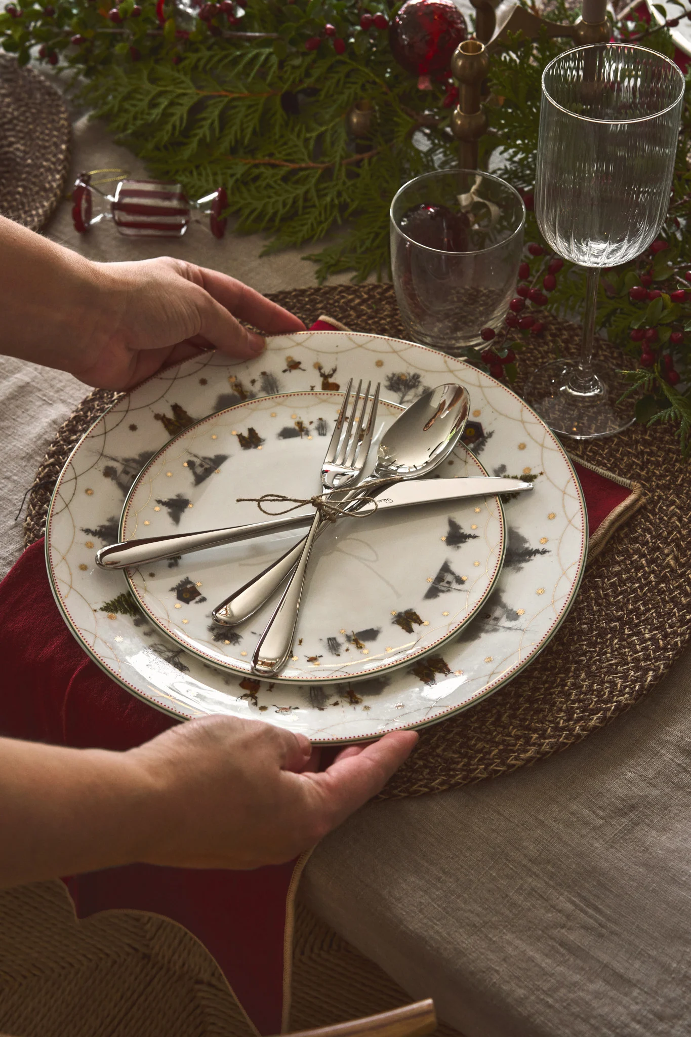A person's hands place a festive dinner plate with a winter forest and reindeer design on a table set with silverware, a red napkin, and greenery.