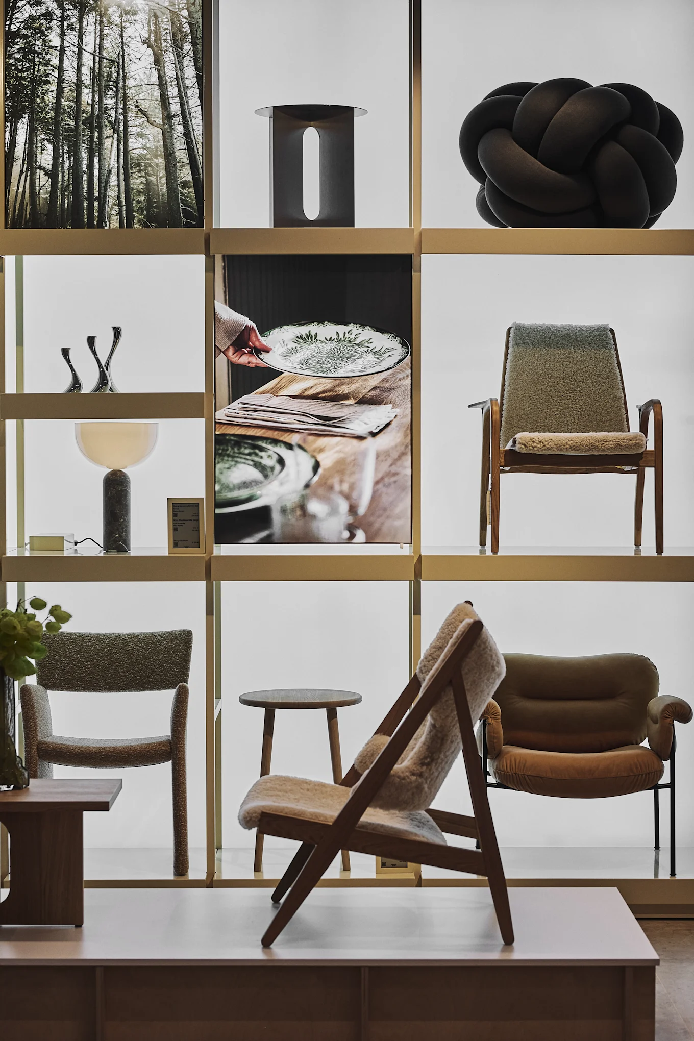 A display with shelves showcasing home decor: a forest print, black knot sculpture, metal art, a table lamp, and various chairs.