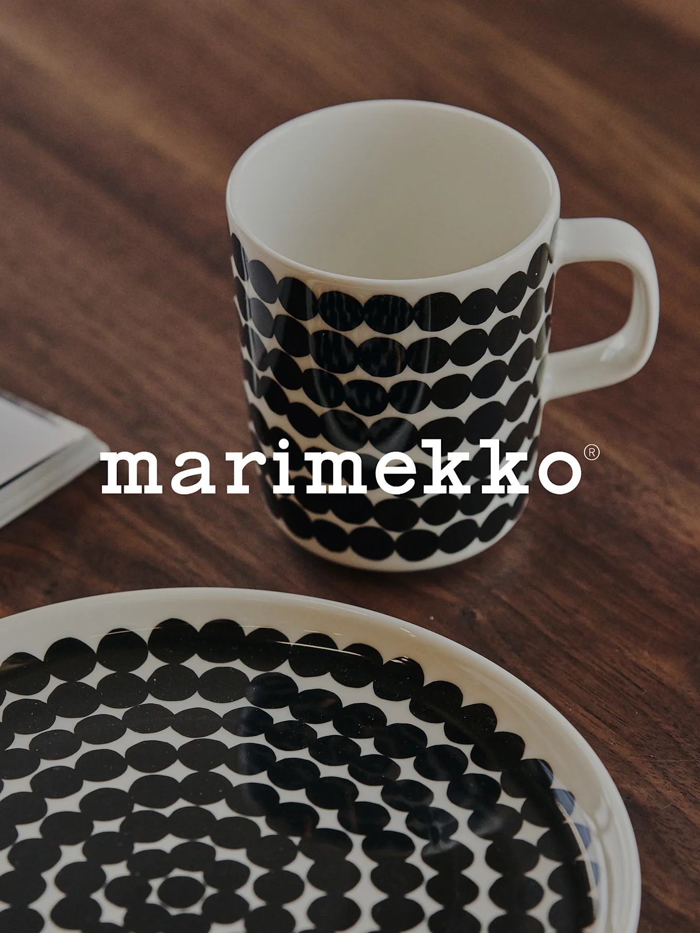 A white mug and plate with a black polka dot pattern on a wooden surface, with the Marimekko logo.