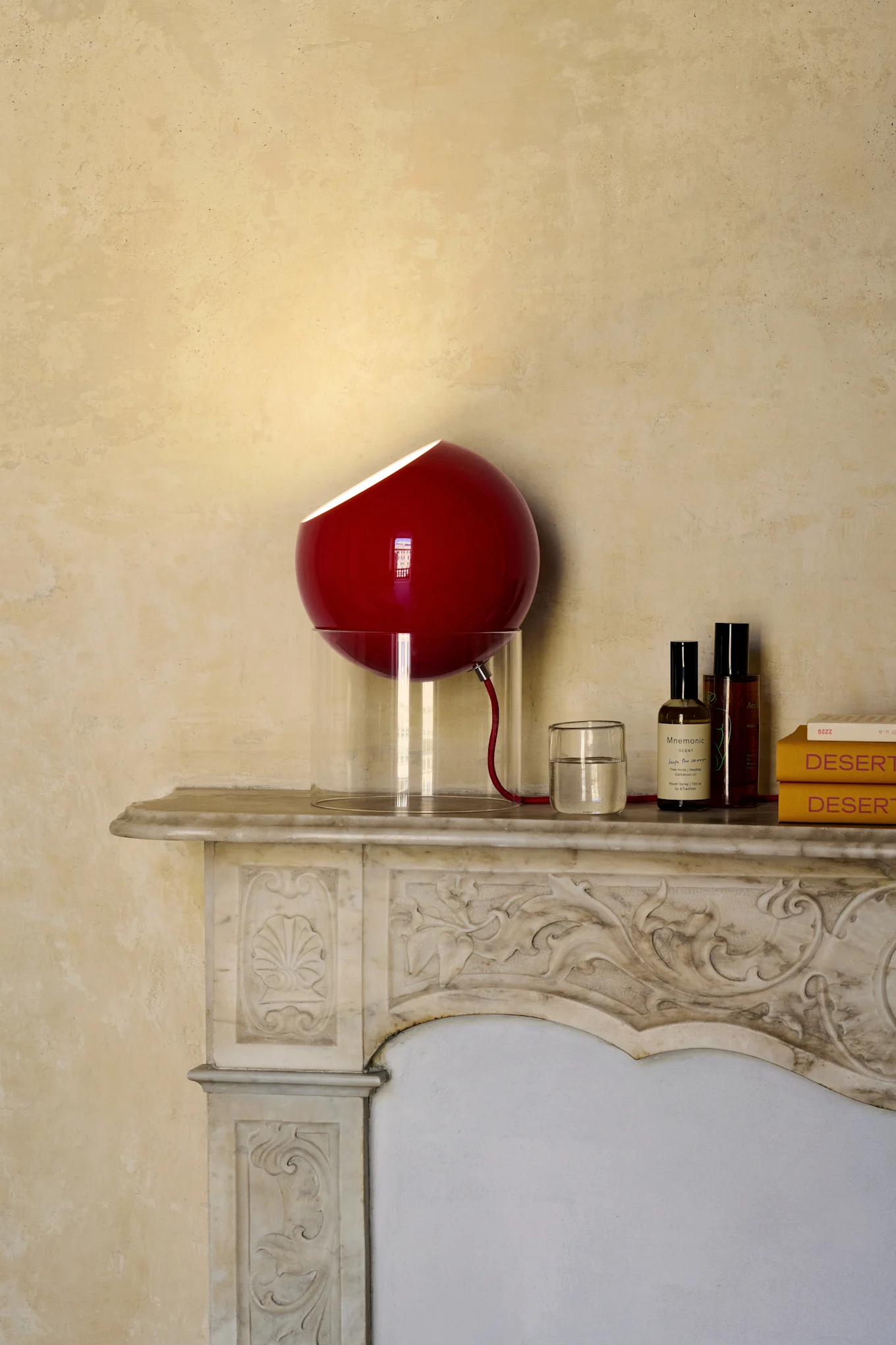 A red spherical lamp with a translucent base sits on a decorative marble mantelpiece next to a glass of water, two bottles, and two yellow books.