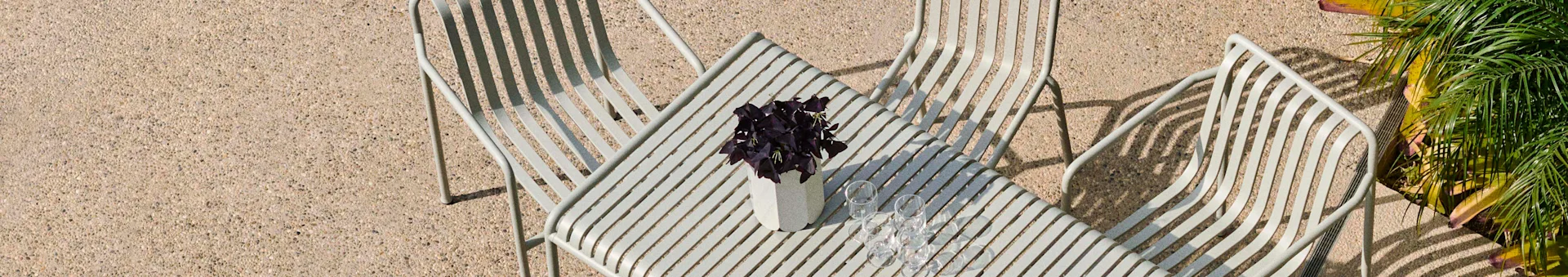 An overhead view of a patio with light grey slatted metal chairs and a matching table holding a purple plant in a white vase and four glasses.