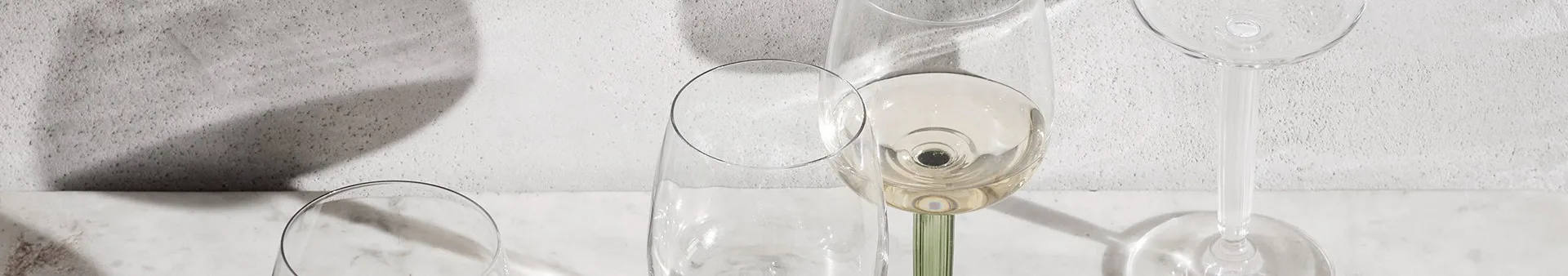 Clear wine glasses on white marble, one with white wine and a green ribbed stem, against a textured gray wall with shadows.