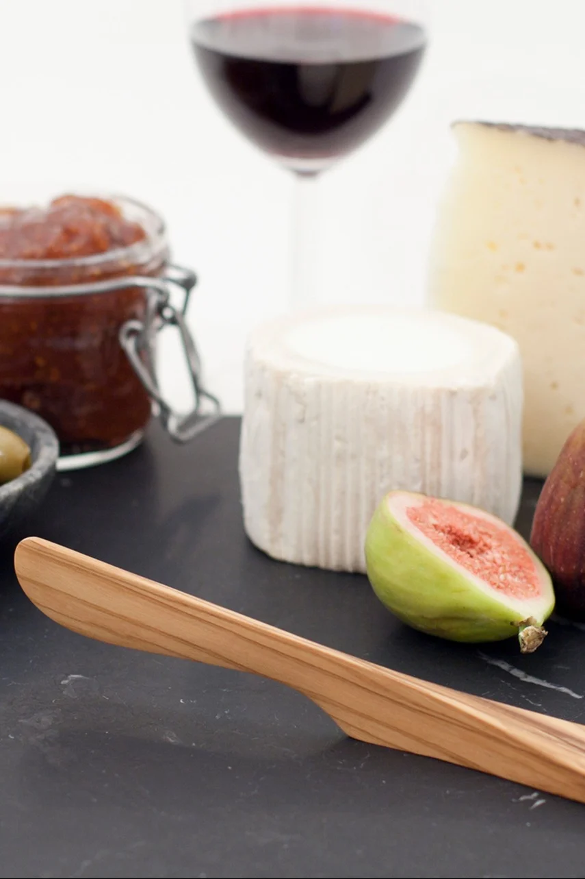 A cheese board with a wooden knife, two cheeses, a halved fig, jam, olives, and a glass of red wine.