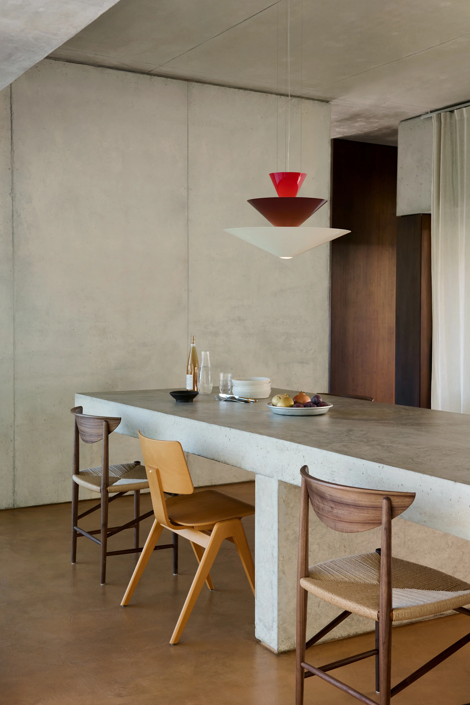 Modern dining area with a concrete island, three wooden chairs, and a tiered red and white pendant light.