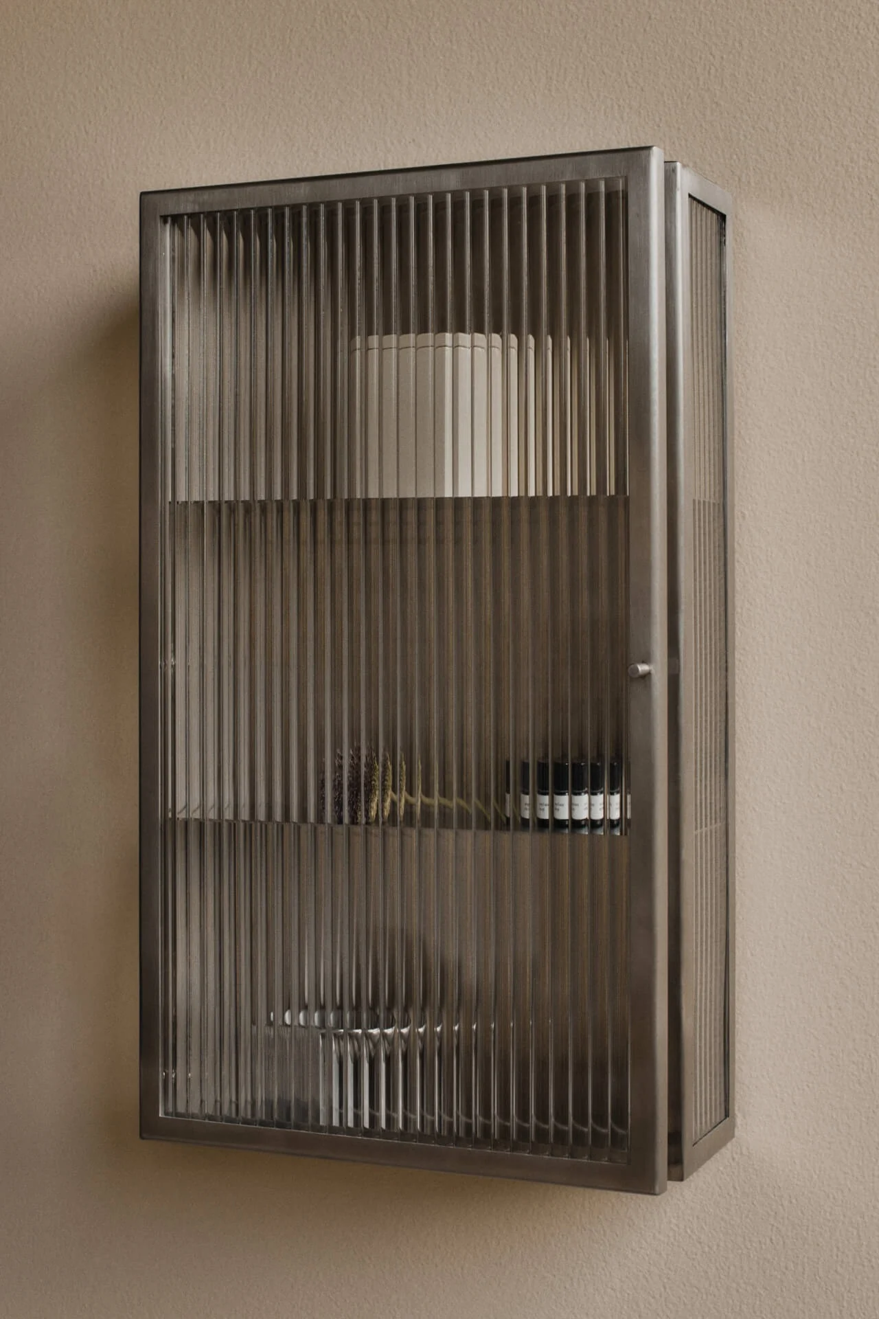 Wall-mounted, dark metal cabinet with fluted glass doors on a beige wall. Contains shelves with a white cylinder, dried botanicals, and small dark bottles.