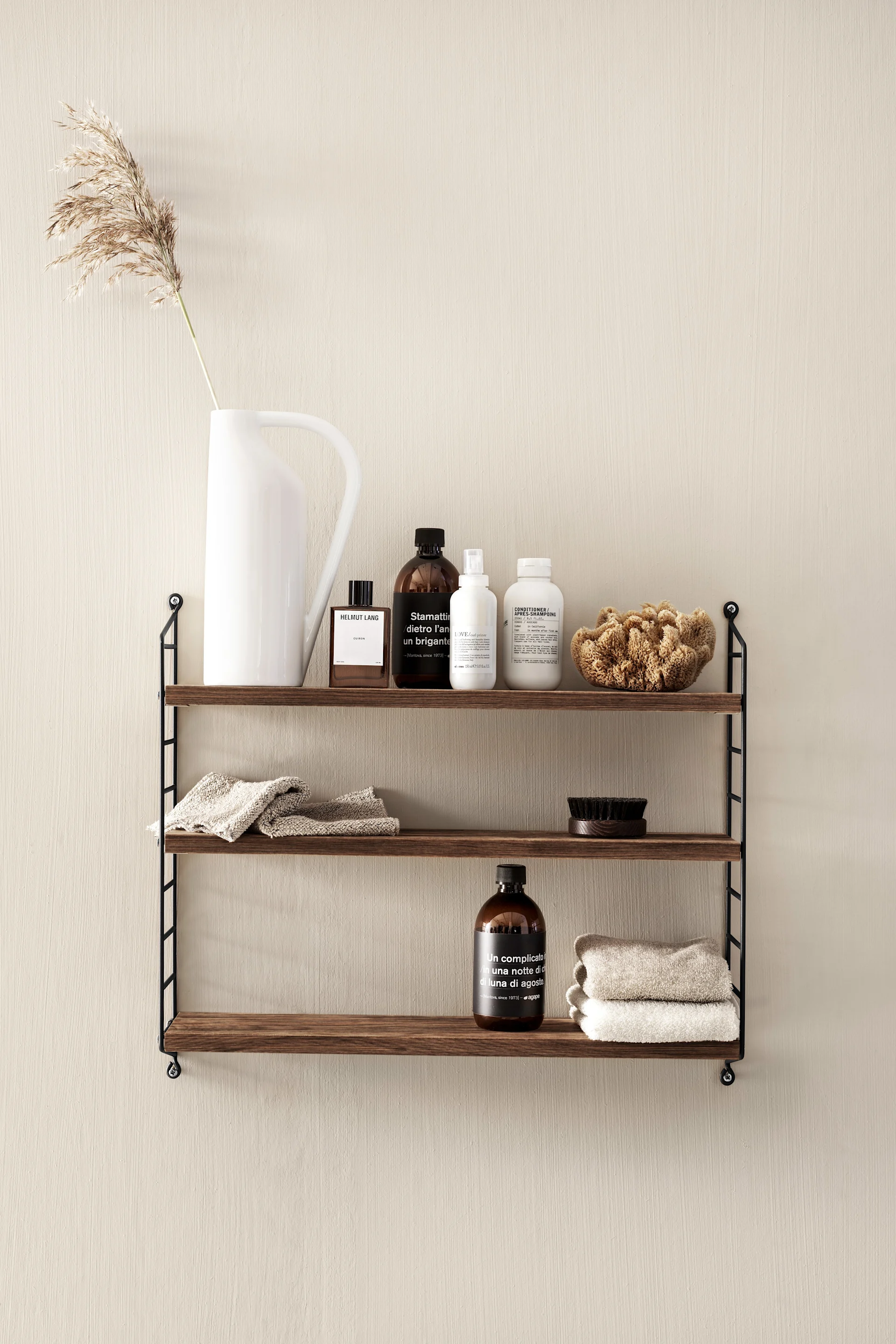 Three-tier dark wood shelf unit on a beige wall with various bottles, folded towels, a brush, and pampas grass.