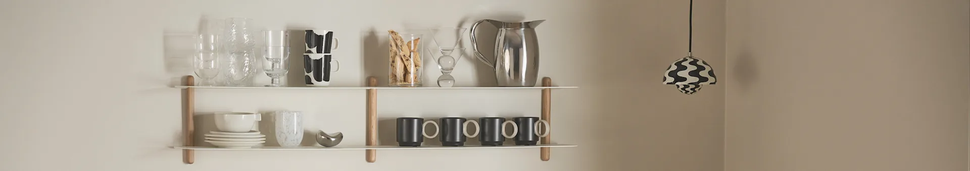 Two white wall shelves with glassware, plates, mugs, and a silver pitcher, with a black and white wavy pendant light on the right.