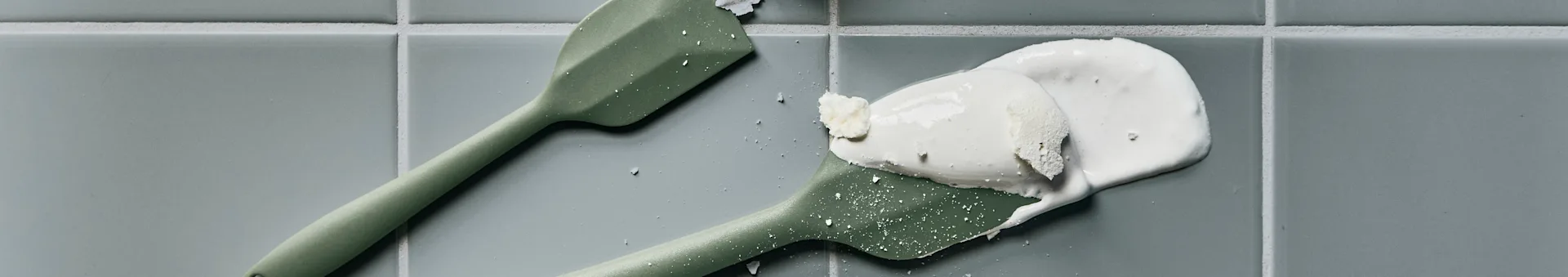 Two green silicone spatulas, one with white cream and flakes, on light gray tiles.
