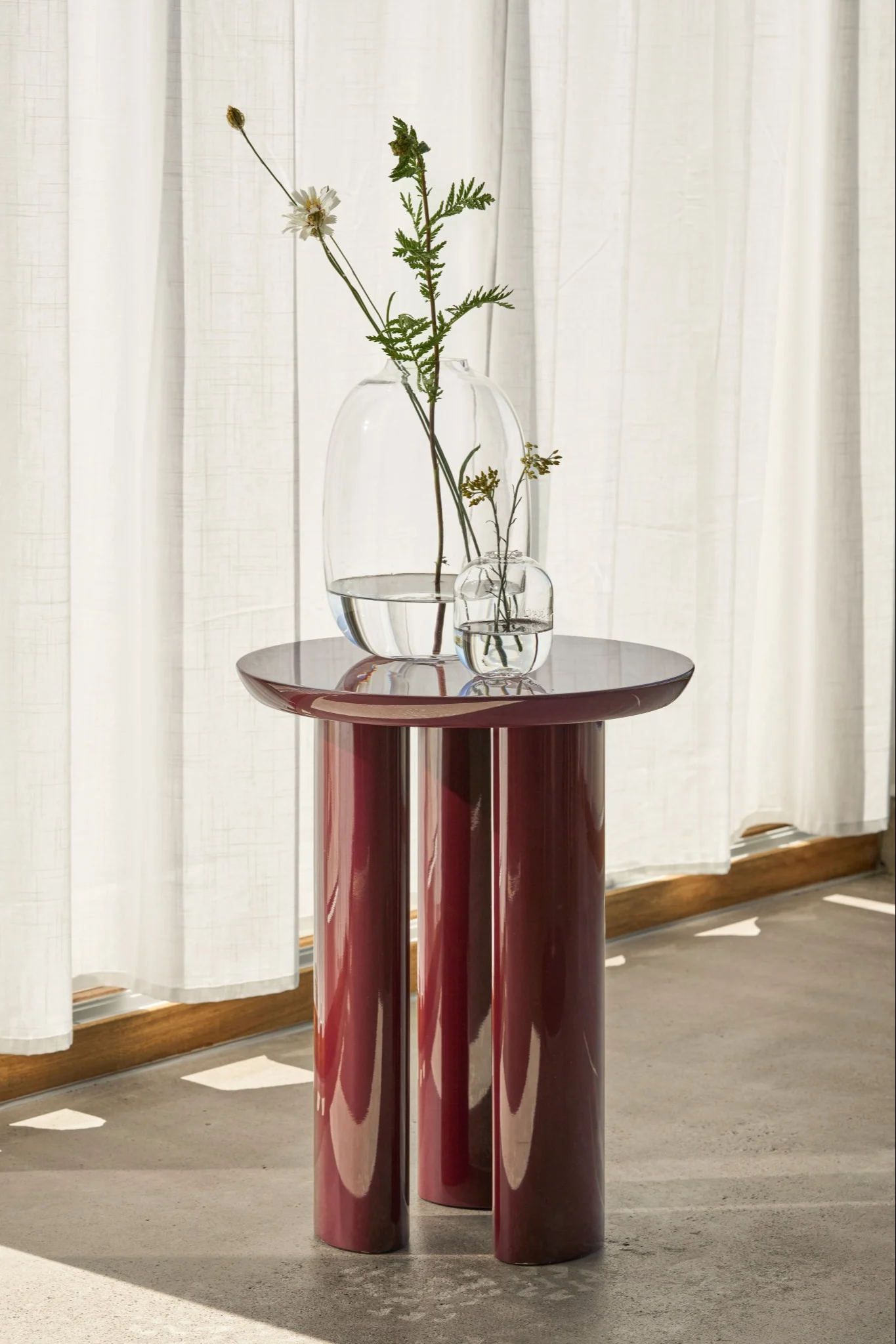 A glossy, round, reddish-brown side table with three cylindrical legs holds two glass vases with a white flower and green foliage.