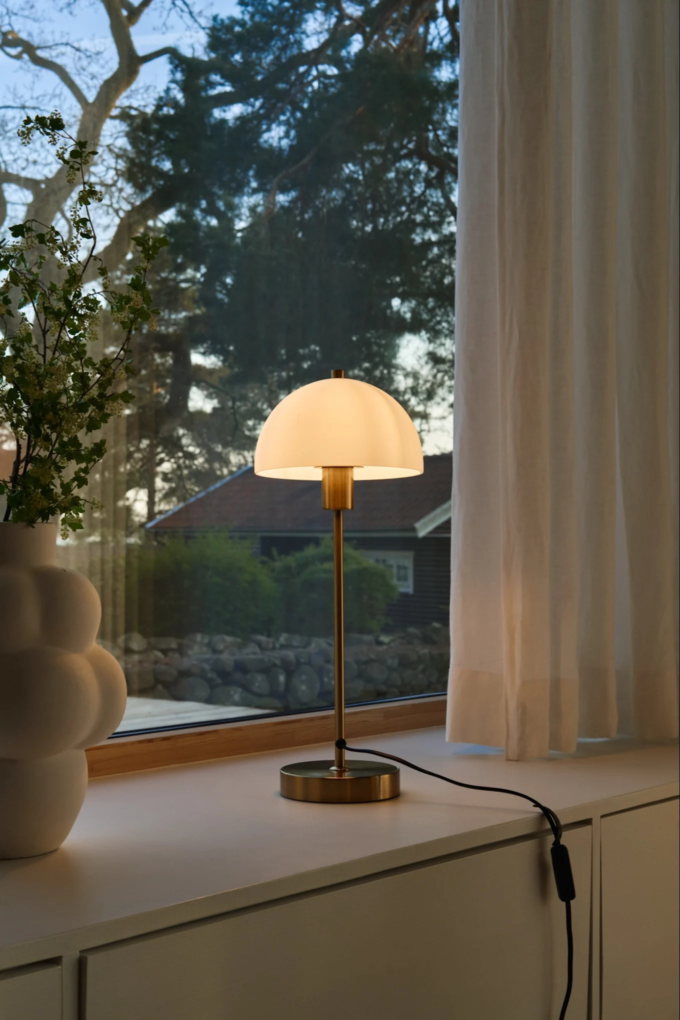 A brass table lamp with a glowing white dome shade sits next to a white sculptural vase with flowering branches on a windowsill.