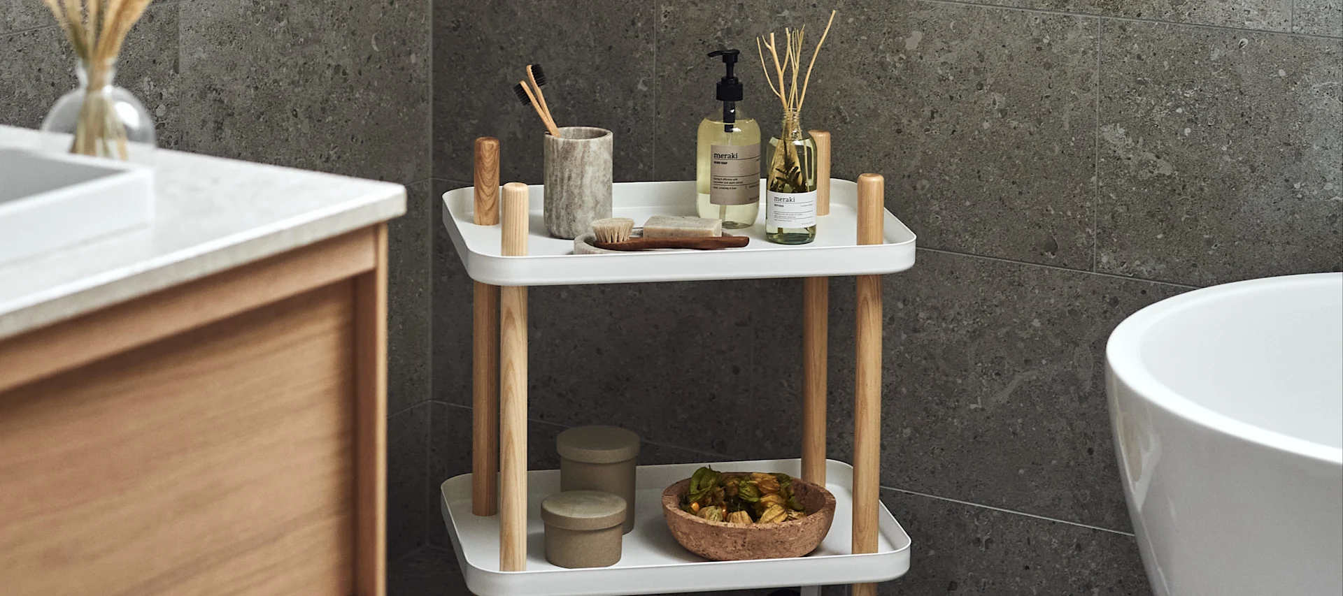 Two-tiered white and wood bathroom caddy holding pump soap, toothbrushes, reed diffuser, and decorative items next to a tub.