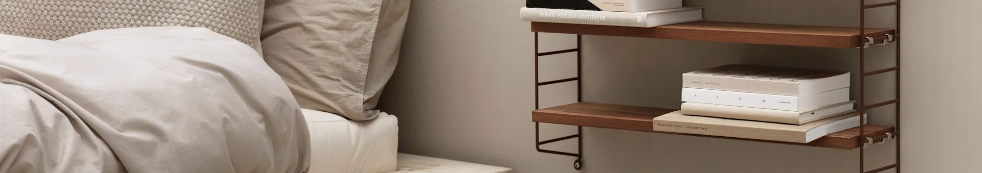 A bed with beige bedding and a knitted headboard, beside a wall-mounted wooden shelving unit holding stacked books.