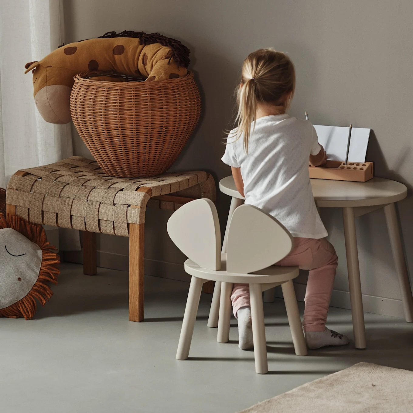 Child drawing at a small round table, seated on a bunny-ear chair, with a giraffe toy in a basket and a woven bench nearby.
