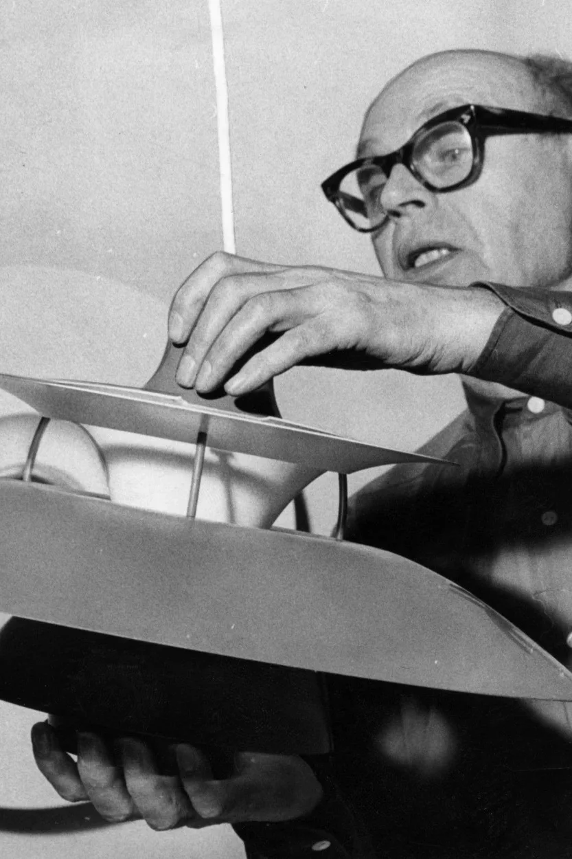 A man in glasses intently adjusts a multi-layered, curved white and gray light fixture or model.