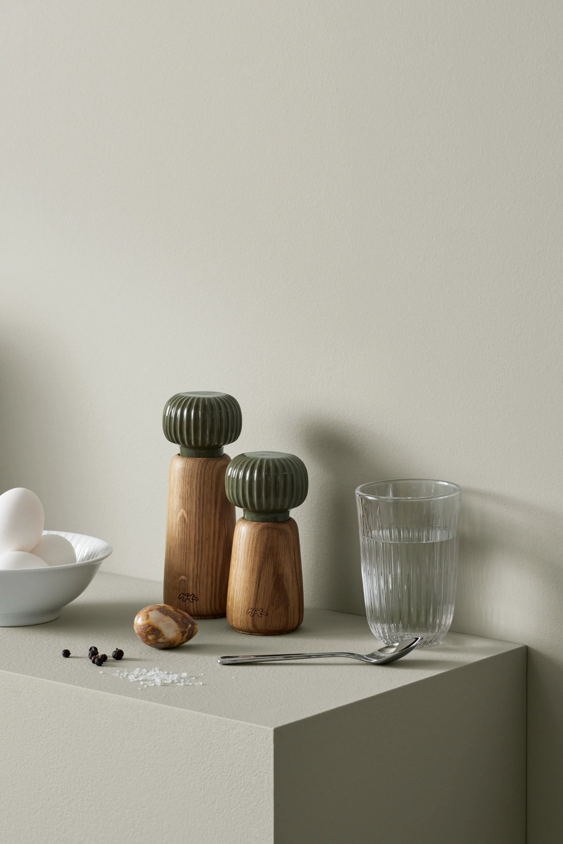Wooden salt and pepper grinders with green tops, eggs in a bowl, a glass of water, and spices on a light grey surface.