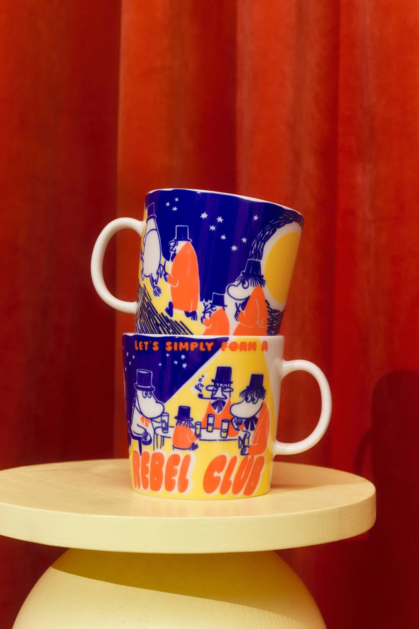 Two stacked mugs, dark blue with yellow and orange Moomin-style characters and 'LET'S SIMPLY FORM A REBEL CLUB', on a yellow table.