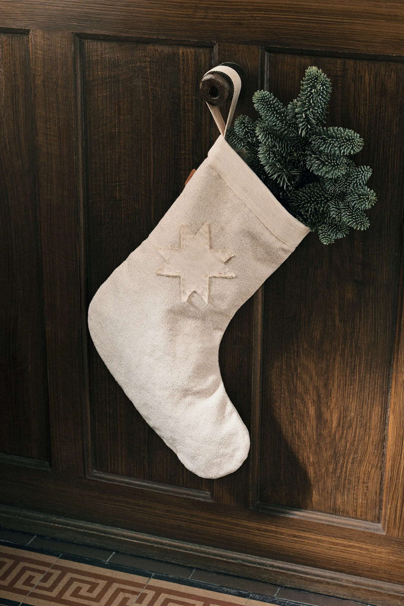 A cream-colored Christmas stocking with a sewn star, filled with pine branches, hangs on a dark wooden door.