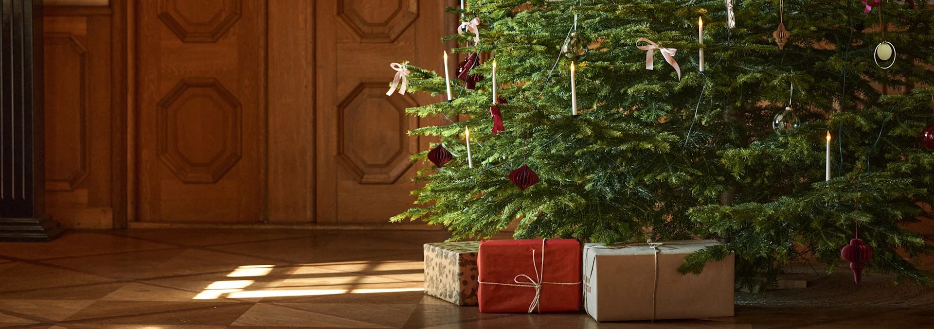 A decorated Christmas tree with glowing candles, pink bows, and paper ornaments, surrounded by three wrapped gifts on a wooden floor with sunlight.