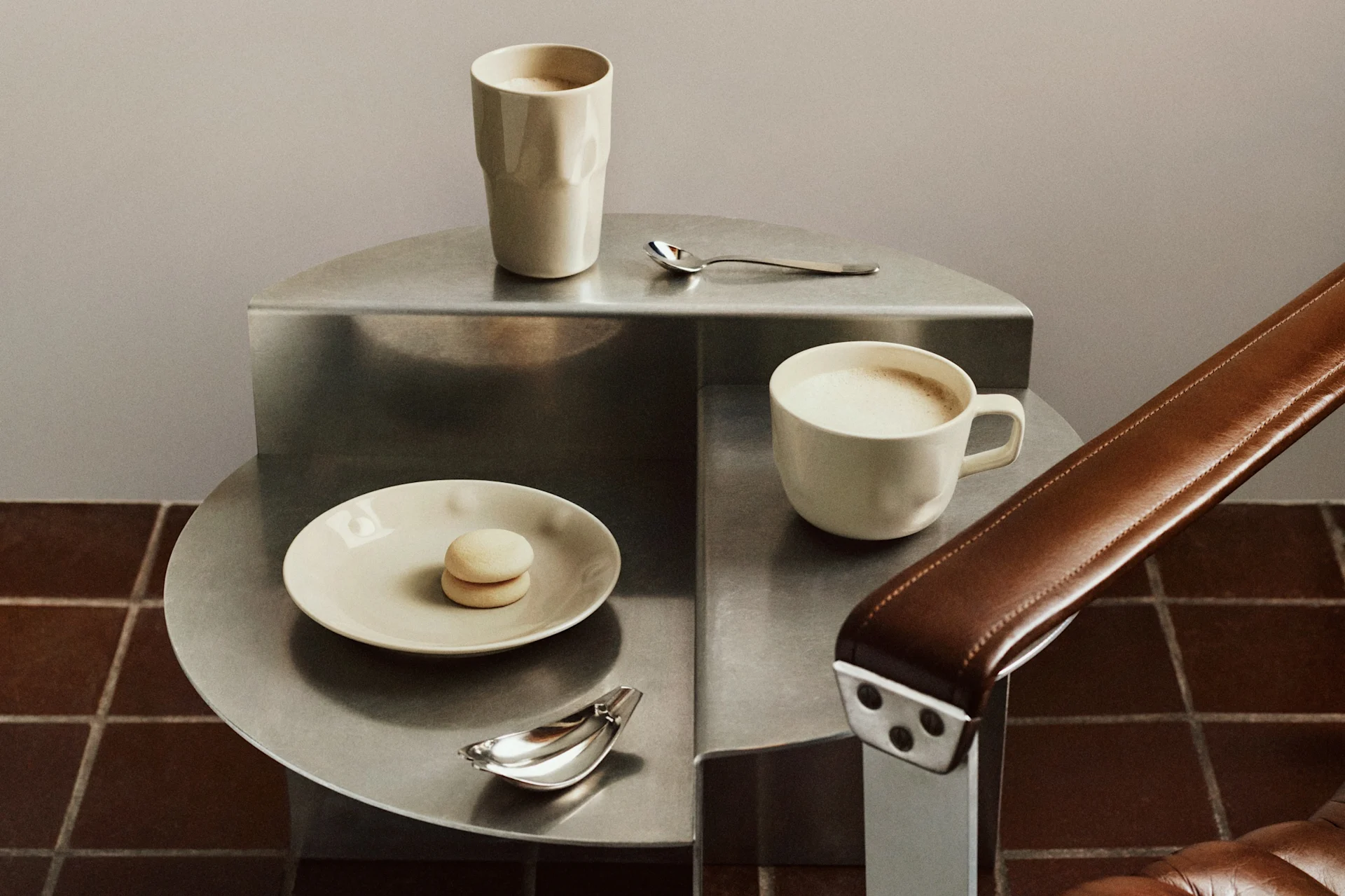 Two light-colored coffee mugs, a macaron, and spoons on a tiered metal side table next to a brown leather armchair.