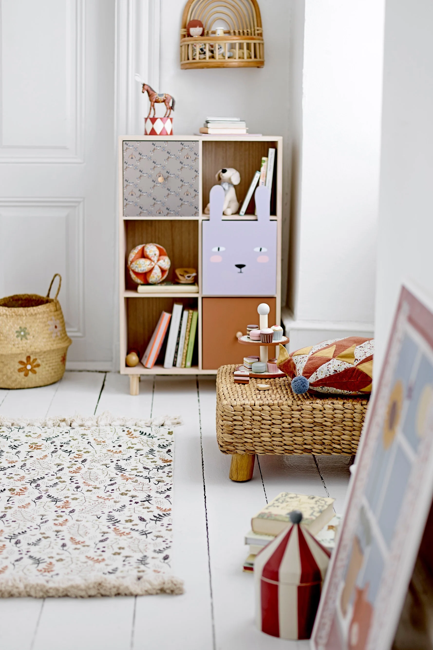 A light-colored child's room with a cubby shelf, woven basket, patterned rug, and toy desserts on a stool.