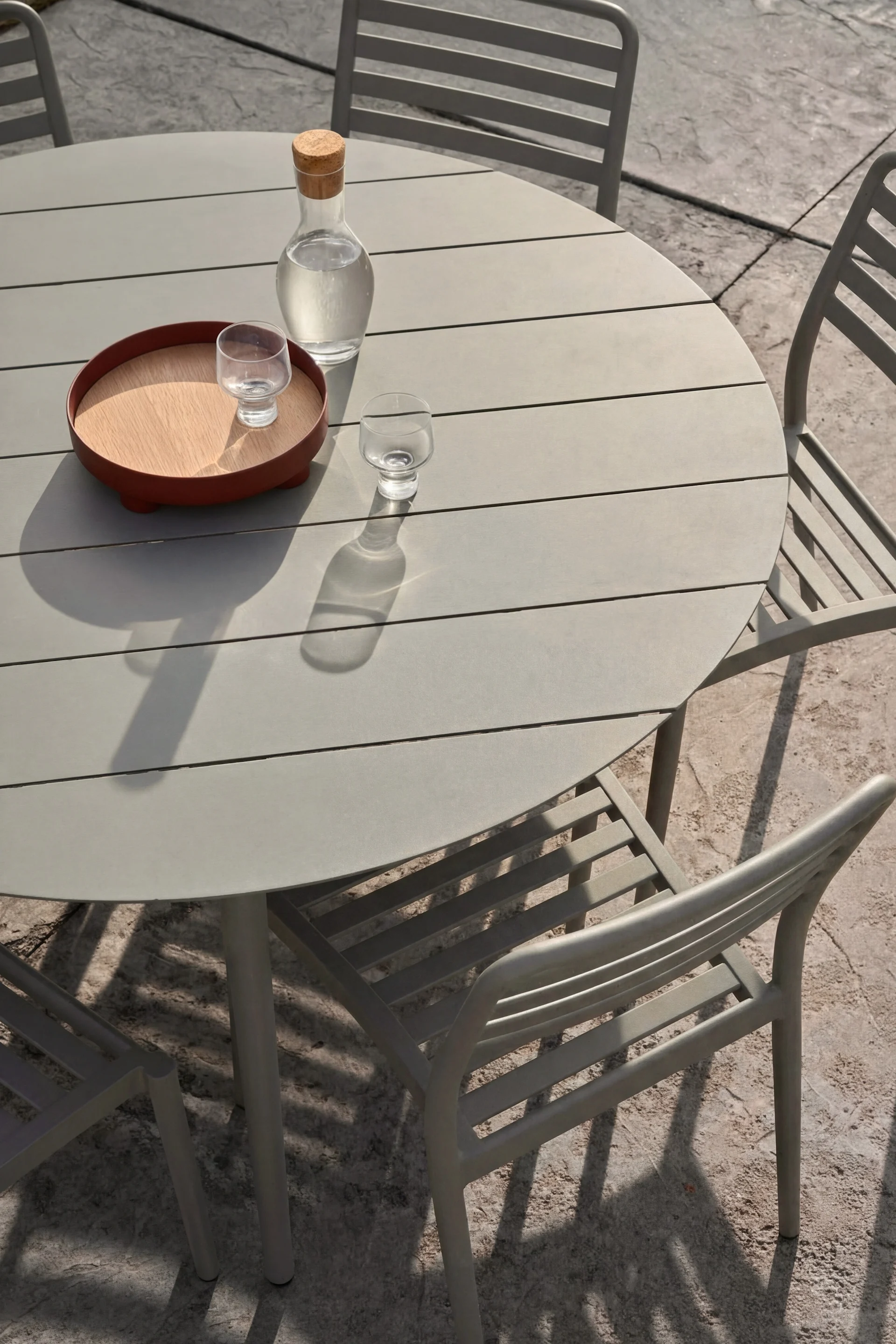 Round grey outdoor table and three chairs. A glass carafe of water and two glasses are on the table, one on a wooden tray.
