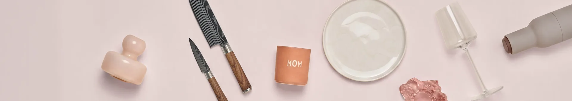 Pink ceramic, two knives, 'MOM' cup, white plate, pink glass, wine glass, and gray bottle on pink surface.
