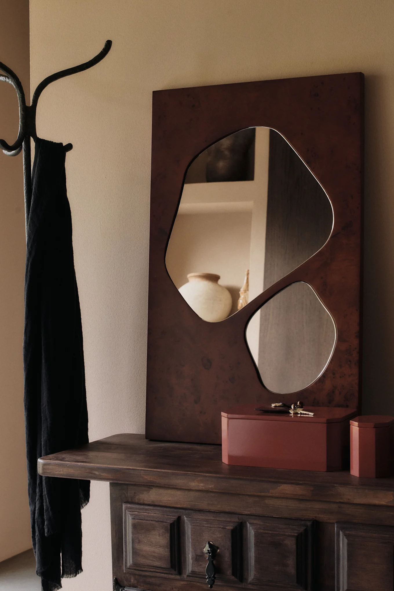 An organic-shaped mirror and two reddish-brown boxes on a wooden dresser next to a coat rack with a black scarf.