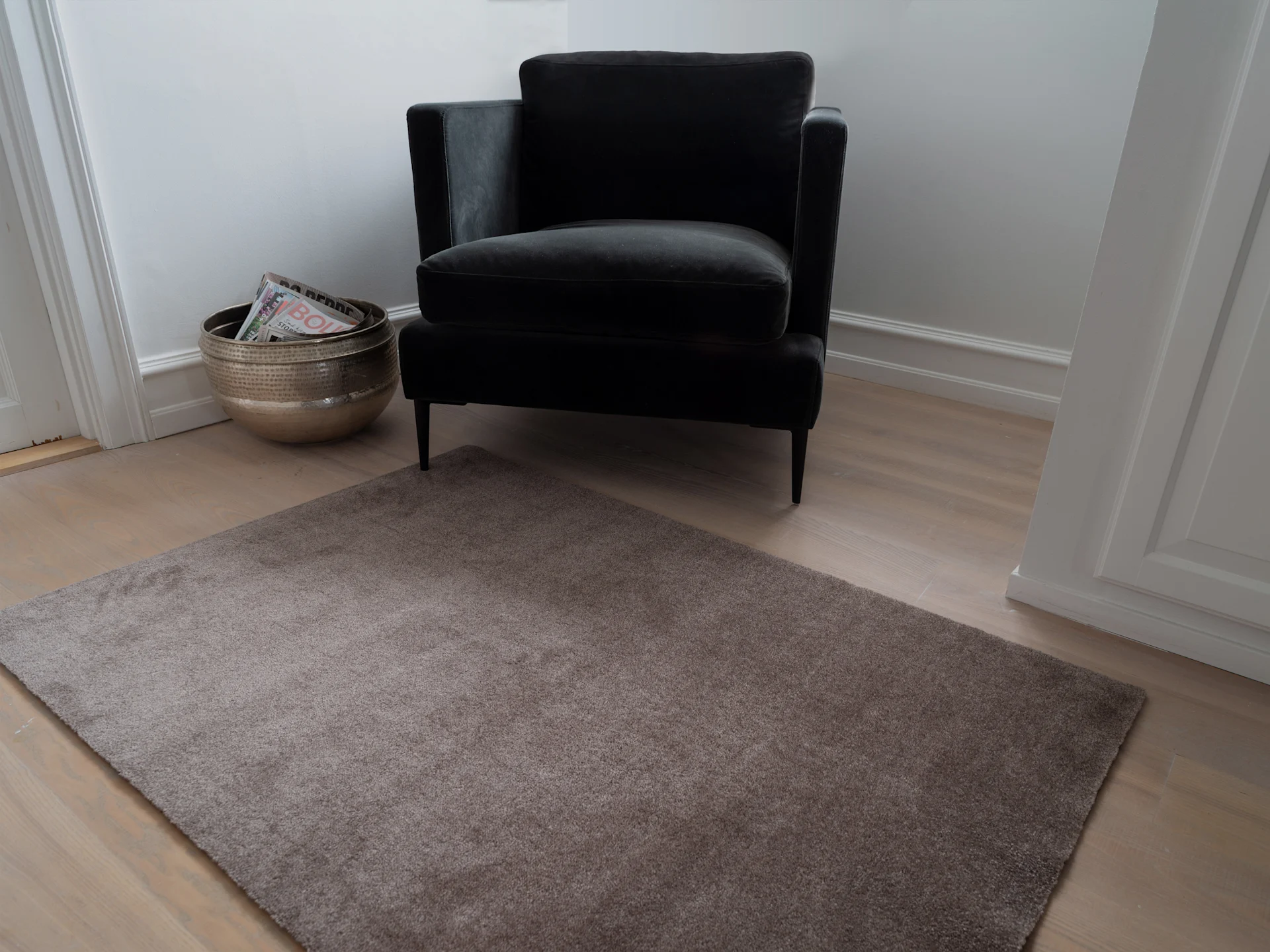 A dark velvet armchair, a golden magazine basket, and a light brown rug on a wooden floor.