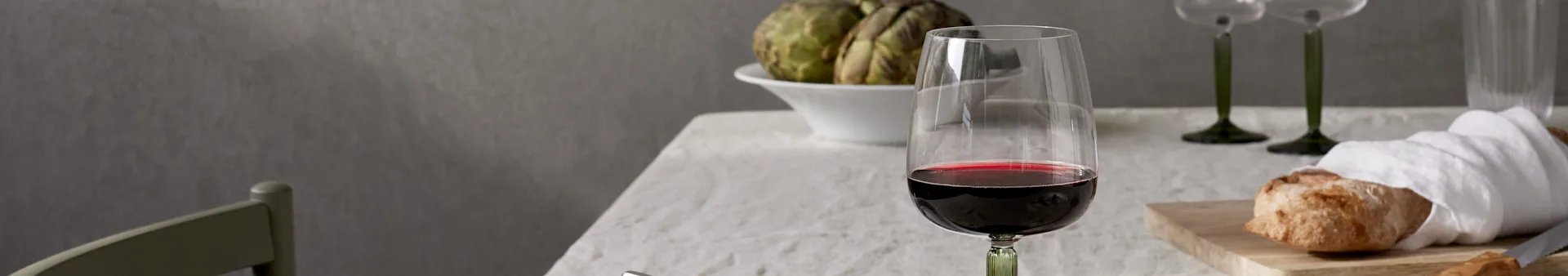 A glass of red wine, artichokes in a bowl, and bread on a cutting board set on a table with a white tablecloth.