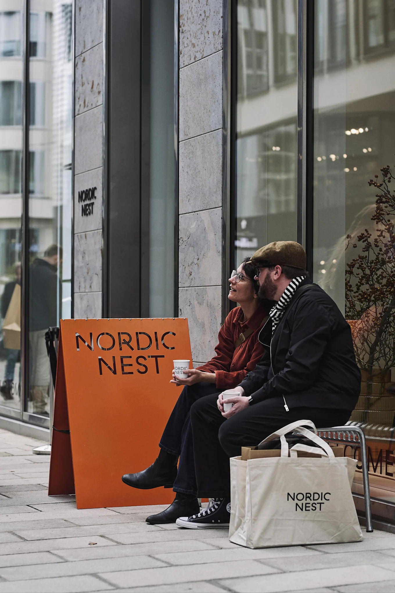 Two people sit outside a store, holding coffee cups with the 'Nordic Nest' logo. An orange sign and a white tote bag also display the logo.