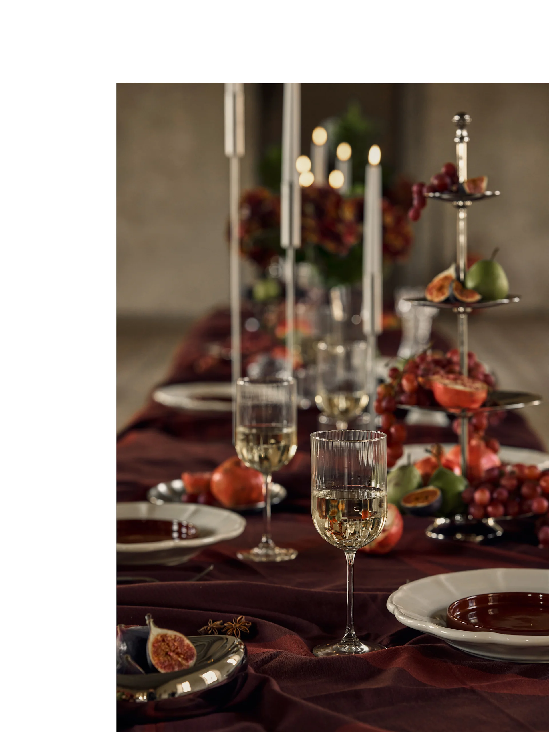 Festive table set with white wine glasses, plates, and a tiered stand filled with colorful fruits and candlelight.