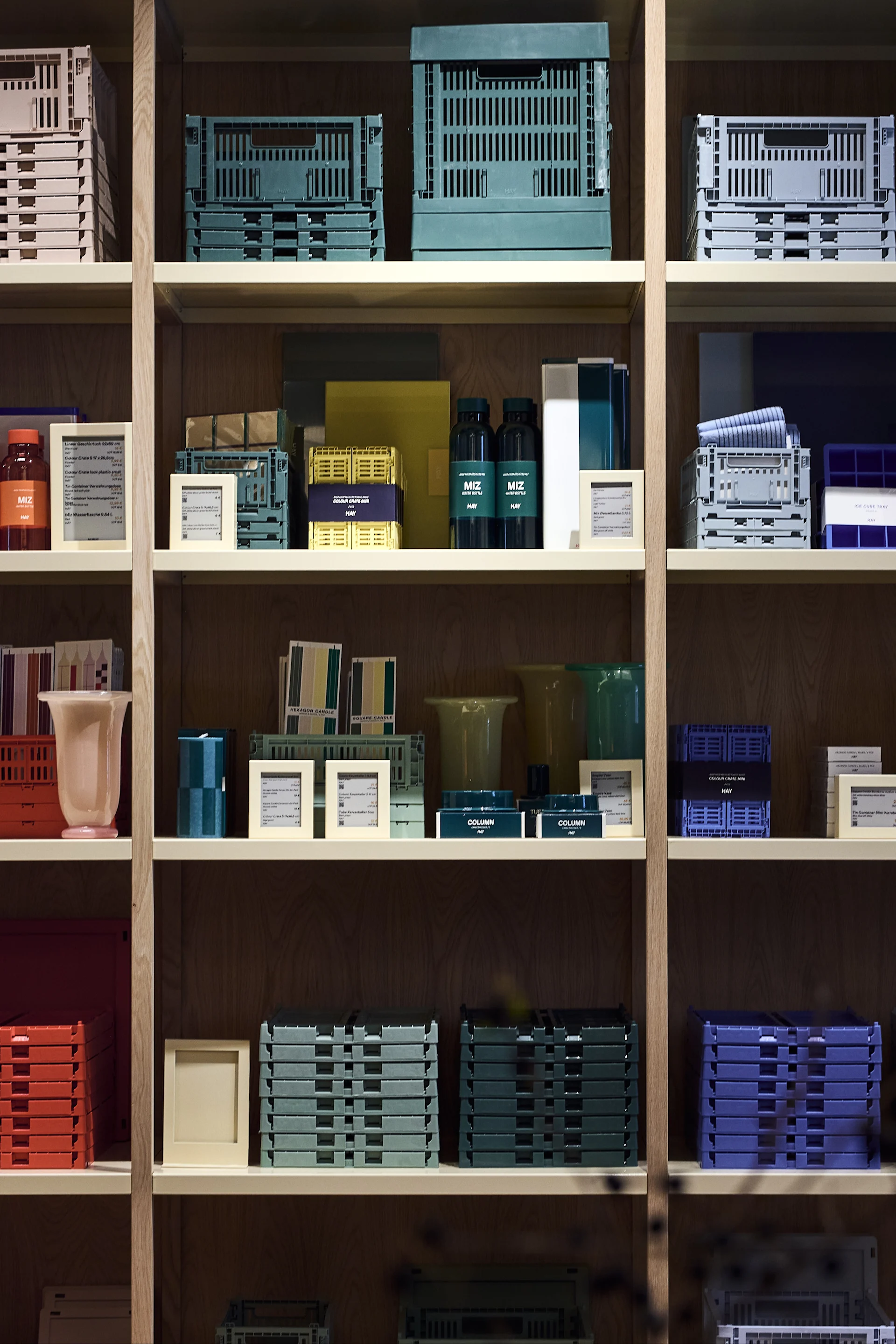 A wooden shelving unit filled with stacks of colorful plastic crates, water bottles, vases, and small display cards.