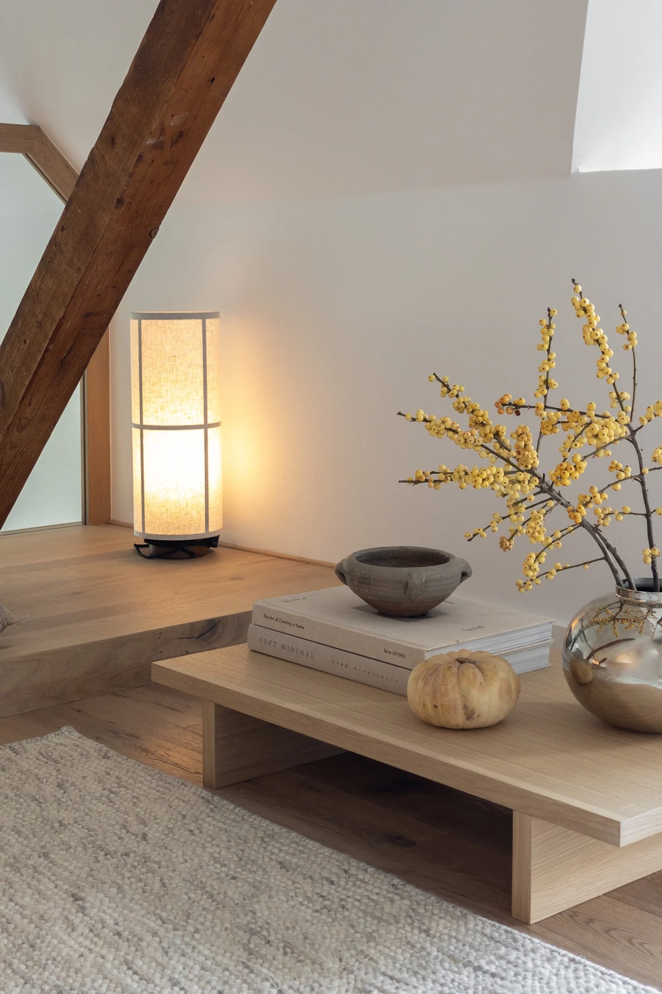 Cozy attic living area with a tall lamp, wooden beam, low coffee table with yellow berries, books, and a small pumpkin.
