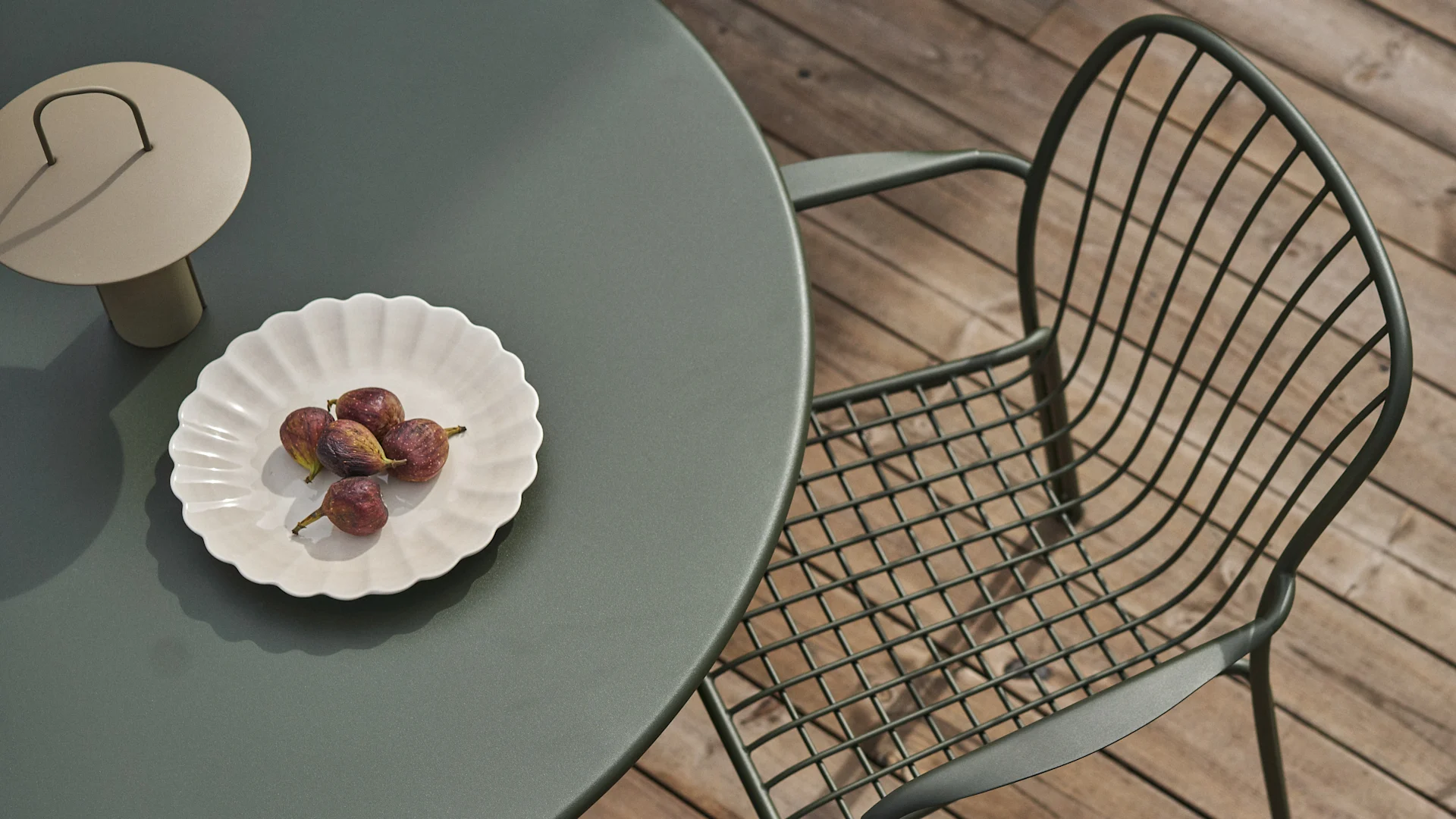 Overhead view of a dark green outdoor table and matching wire chair on a wooden deck. A white scalloped plate with figs and a beige lamp are on the table.