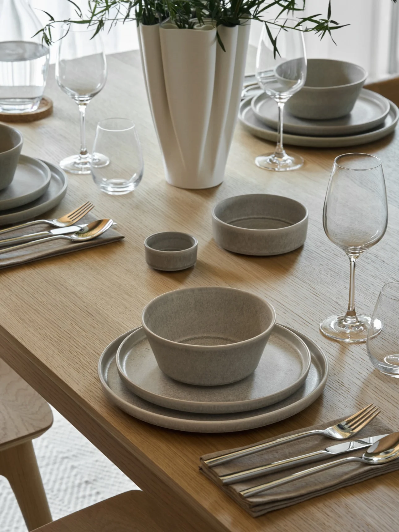 A light wooden dining table set with grey ceramic dishes, silver cutlery, clear glasses, and a white vase with green foliage.