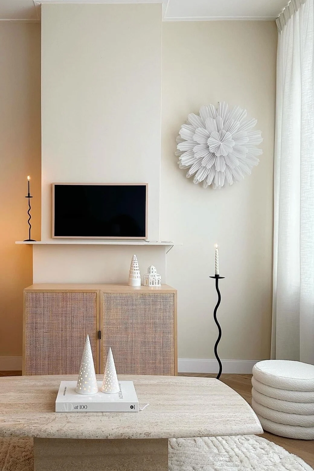 A neutral living room with a textured rug, travertine coffee table, rattan cabinet under a TV, a tall floor lamp, and a white pleated wall decoration.