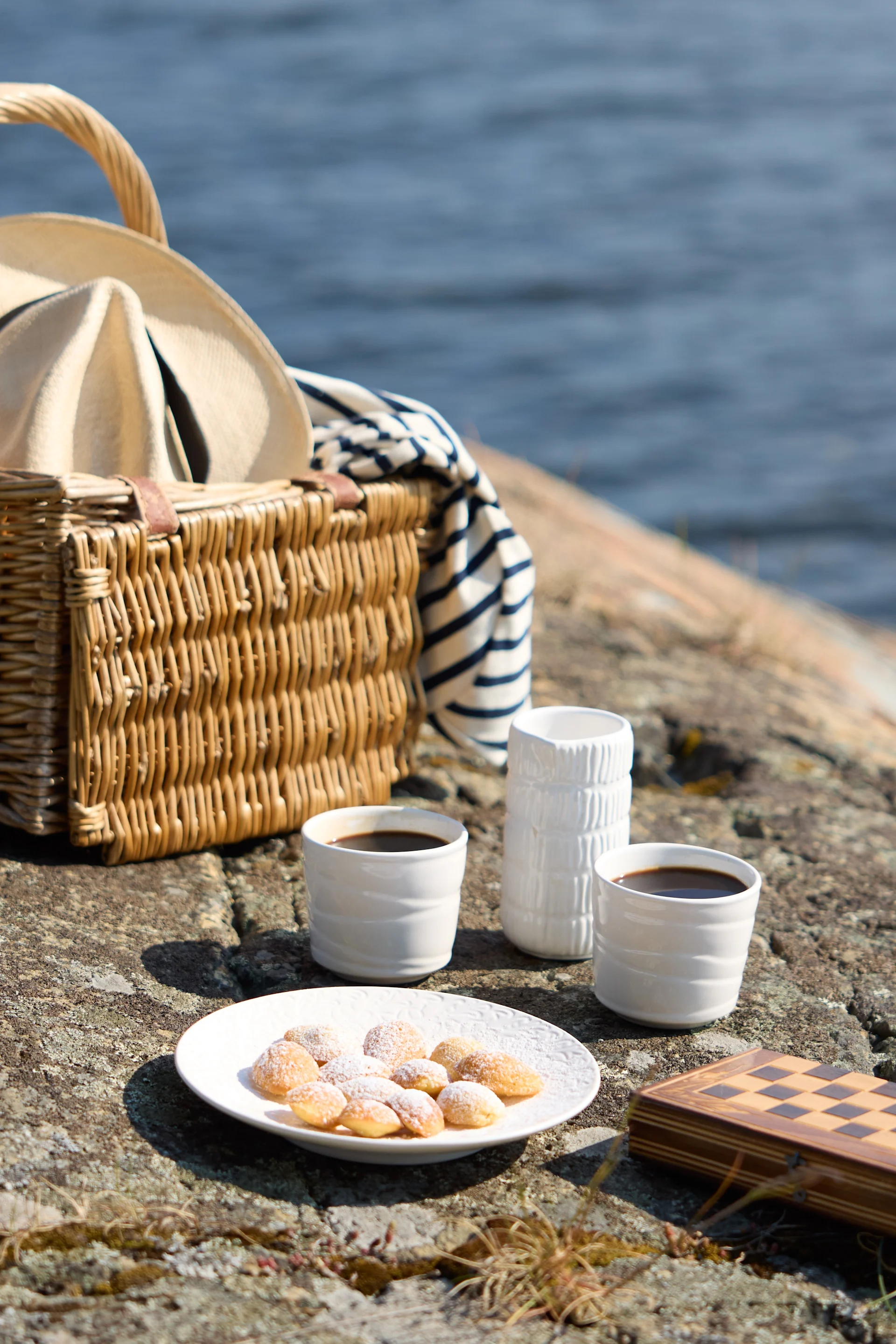 A picnic setting on rocky terrain near water, featuring coffee, pastries, and a chess set.