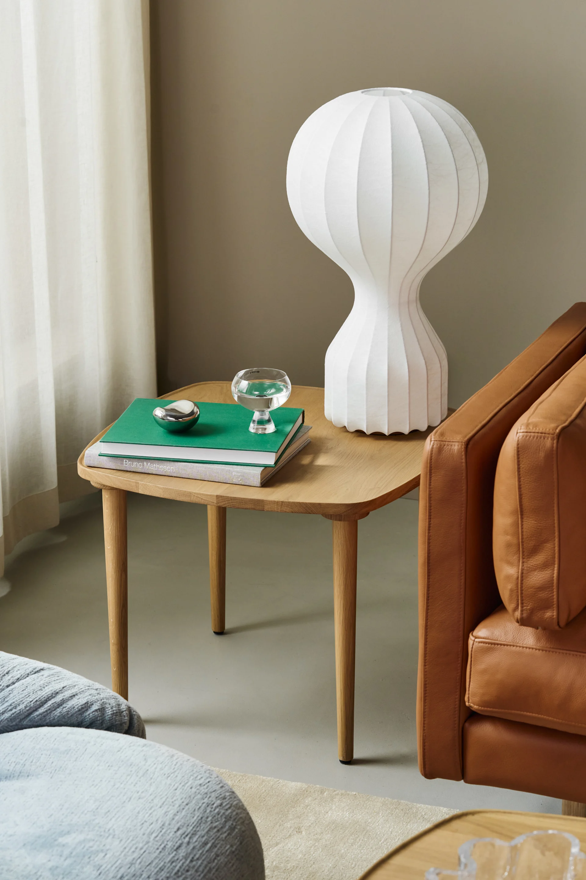 A sculptural white table lamp, books, a glass, and a silver object on a wooden side table next to a brown leather armchair.