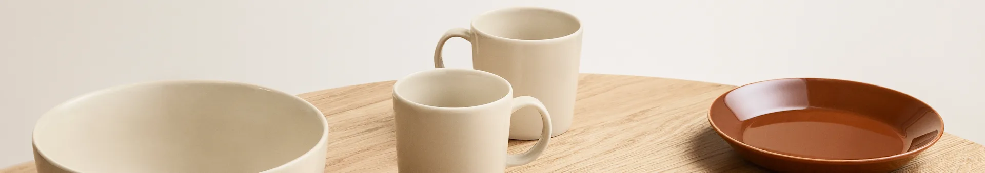 Assortment of light beige ceramic mugs and a reddish-brown bowl on a wooden surface.