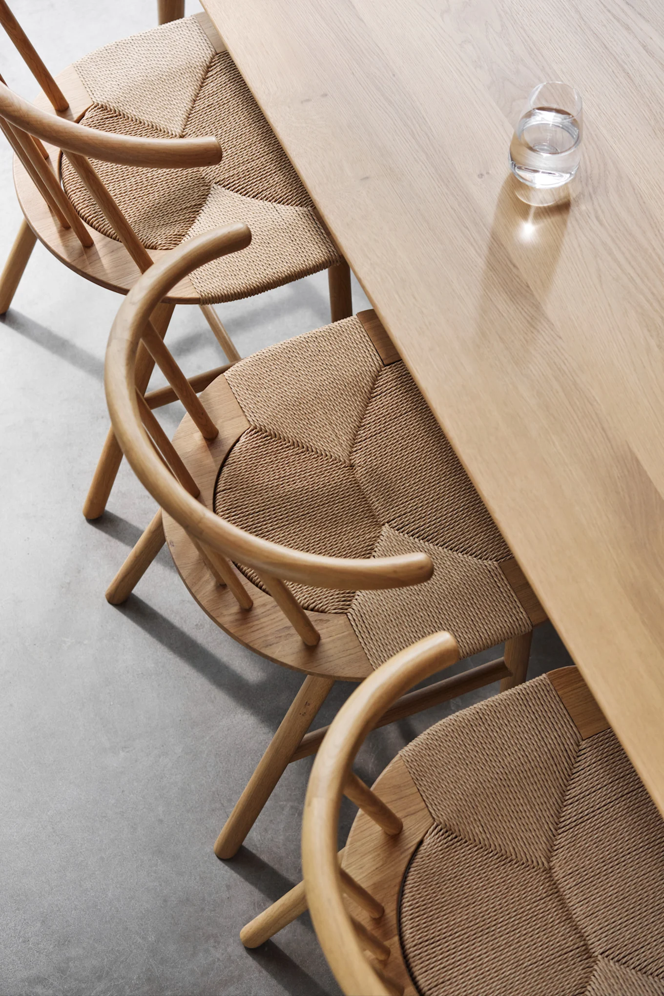 Overhead view of a light wooden dining table with three matching chairs featuring woven rattan seats and a glass of water on the table.