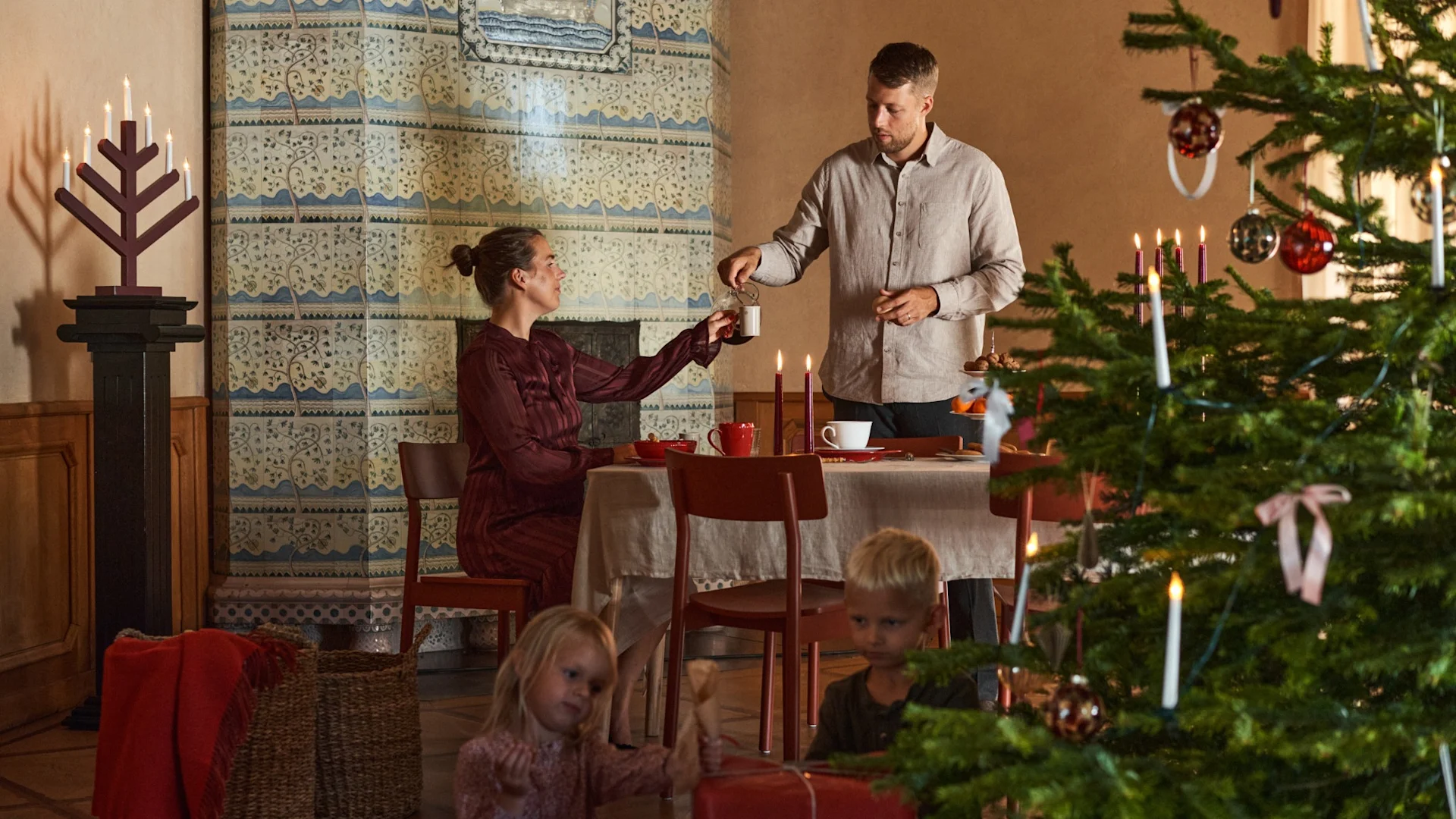 A couple sits at a table decorated for Christmas, next to a Christmas tree with children unwrapping presents.