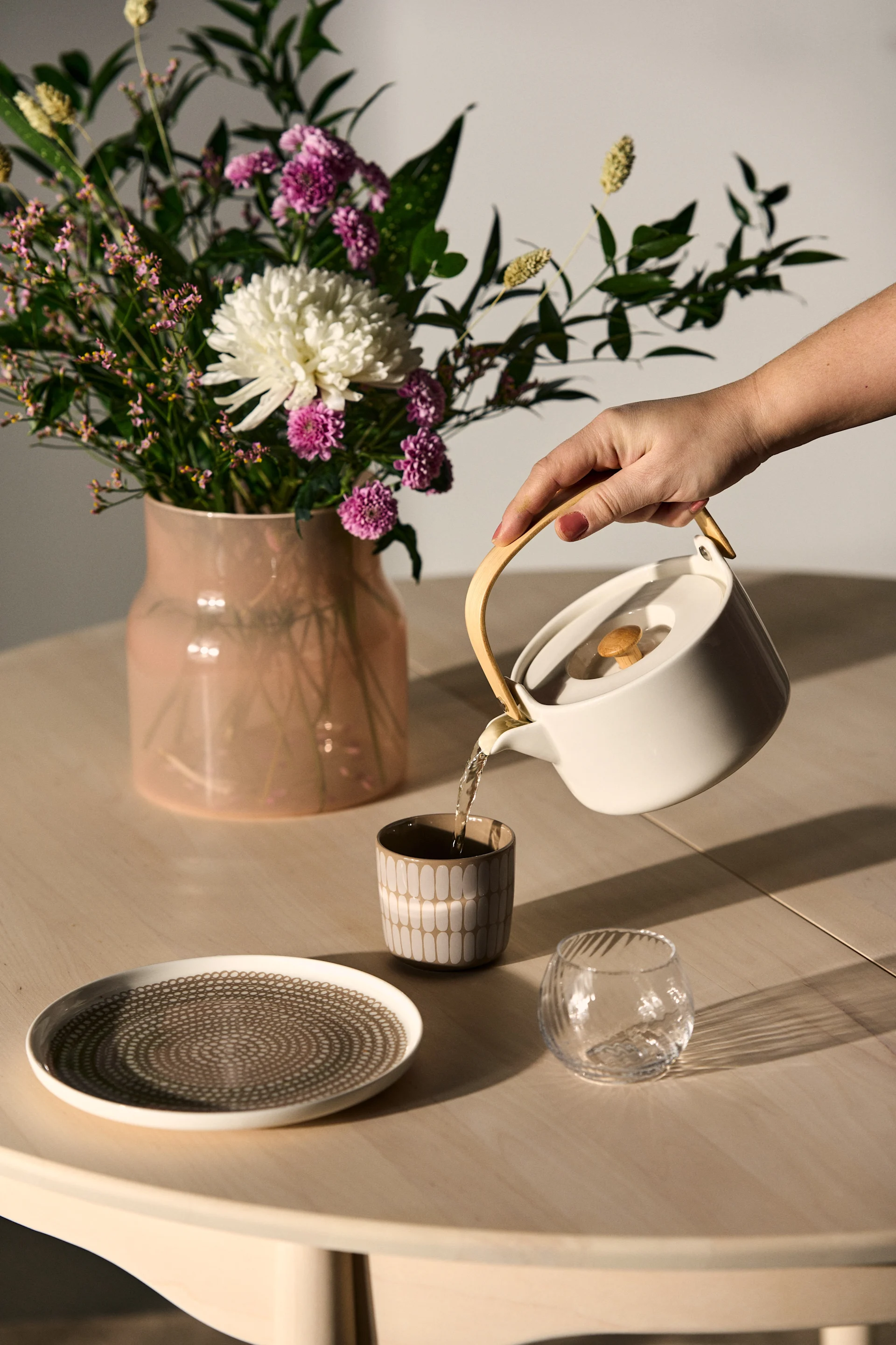 A hand pours from a white teapot with a wooden handle into a beige patterned cup, next to a vase of flowers and a decorative plate.