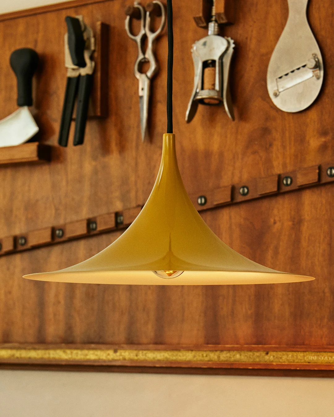 Golden bell-shaped pendant lamp with visible bulb, against a wooden wall with hanging tools.