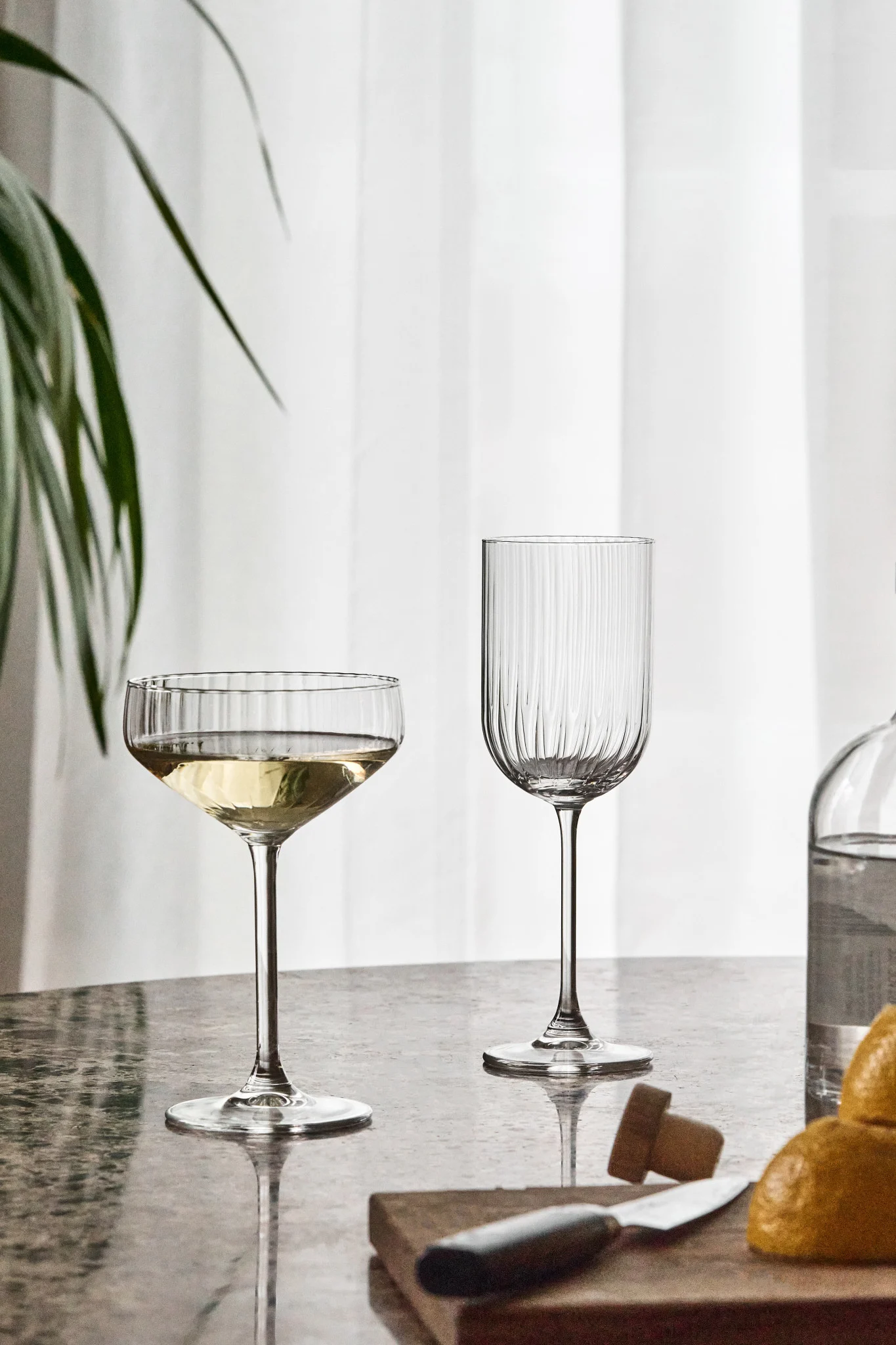 Two fluted glasses on a reflective table, one filled with white liquid. Lemons, a knife, and a bottle are blurred in the foreground.